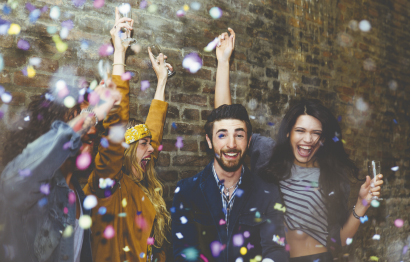 Eine Gruppe von fröhlichen Menschen feiert mit Konfetti und Getränken vor einer Ziegelwand.
